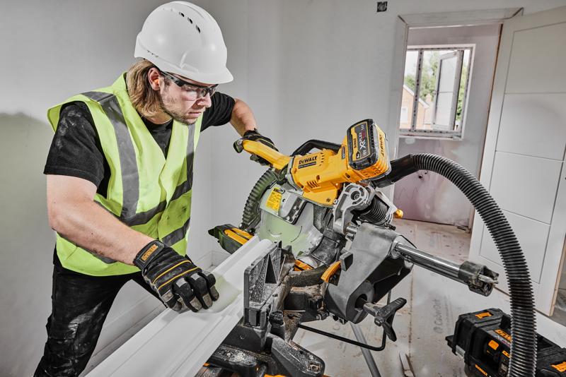 Una persona con chaleco de seguridad, guantes y casco está utilizando una sierra de inglete inalámbrica DEWALT DCS781X2 para cortar una moldura dentro de una habitación en construcción. La sierra está conectada a un sistema de extracción de polvo y se pueden ver baterías de herramientas DEWALT cerca.