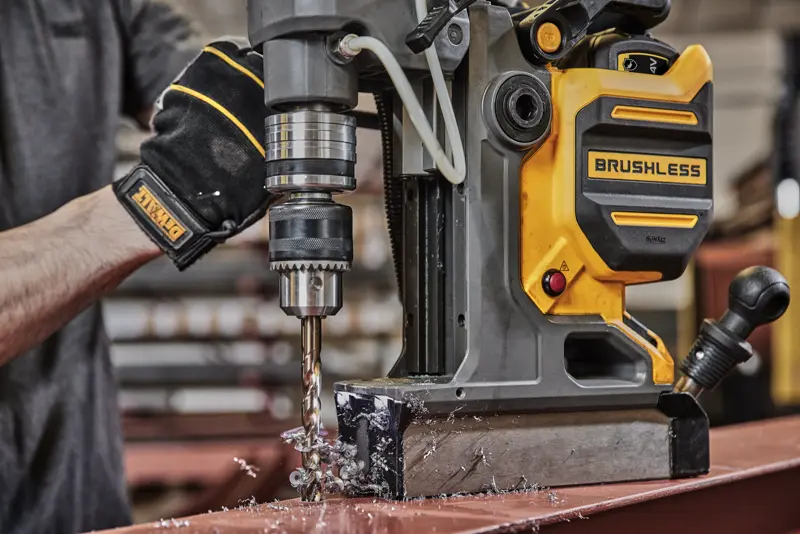 Persona usa taladro magnético DEWALT brushless para perforar metal, creando virutas sobre mesa de taller.