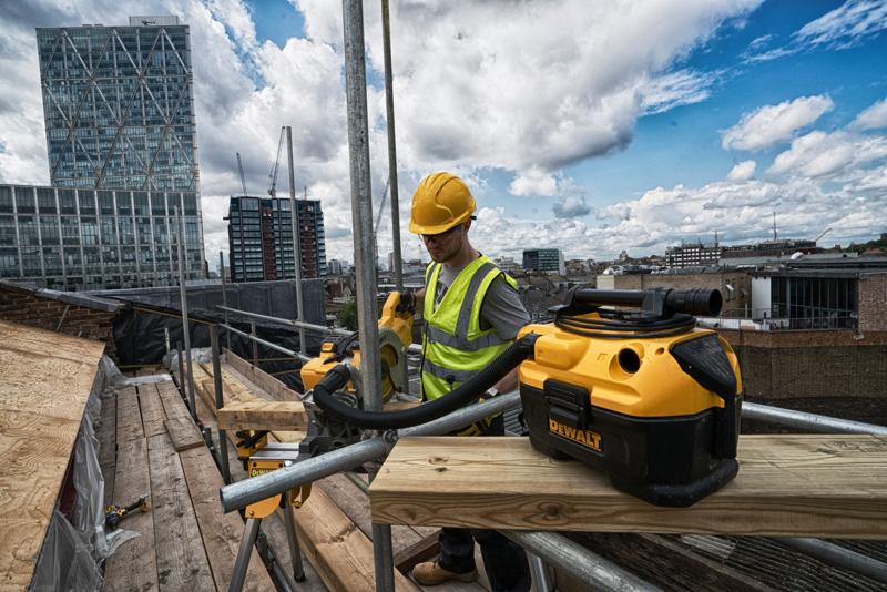 Un aspirador portátil DEWALT DCV584 en una obra de construcción, con un trabajador usando chaleco reflectante y casco operando herramientas eléctricas sobre un andamio, y edificios urbanos visibles en el fondo.