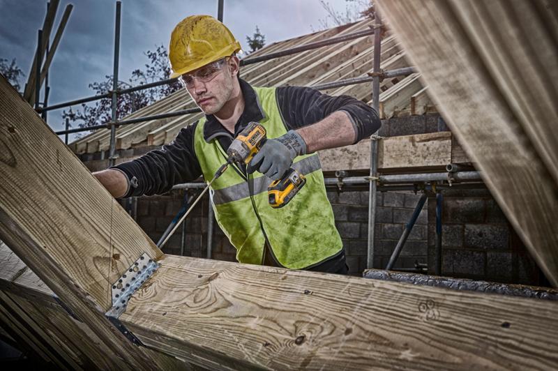 Un trabajador de la construcción con casco amarillo y chaleco reflectante utiliza un taladro inalámbrico DEWALT para fijar soportes metálicos en vigas de madera en una obra. Su rostro está desenfocado.