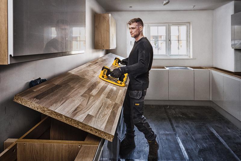 Una persona con el rostro difuminado está instalando una encimera de madera en una cocina usando una herramienta eléctrica DEWALT. La cocina es moderna, con gabinetes de colores claros y una ventana grande en el fondo.