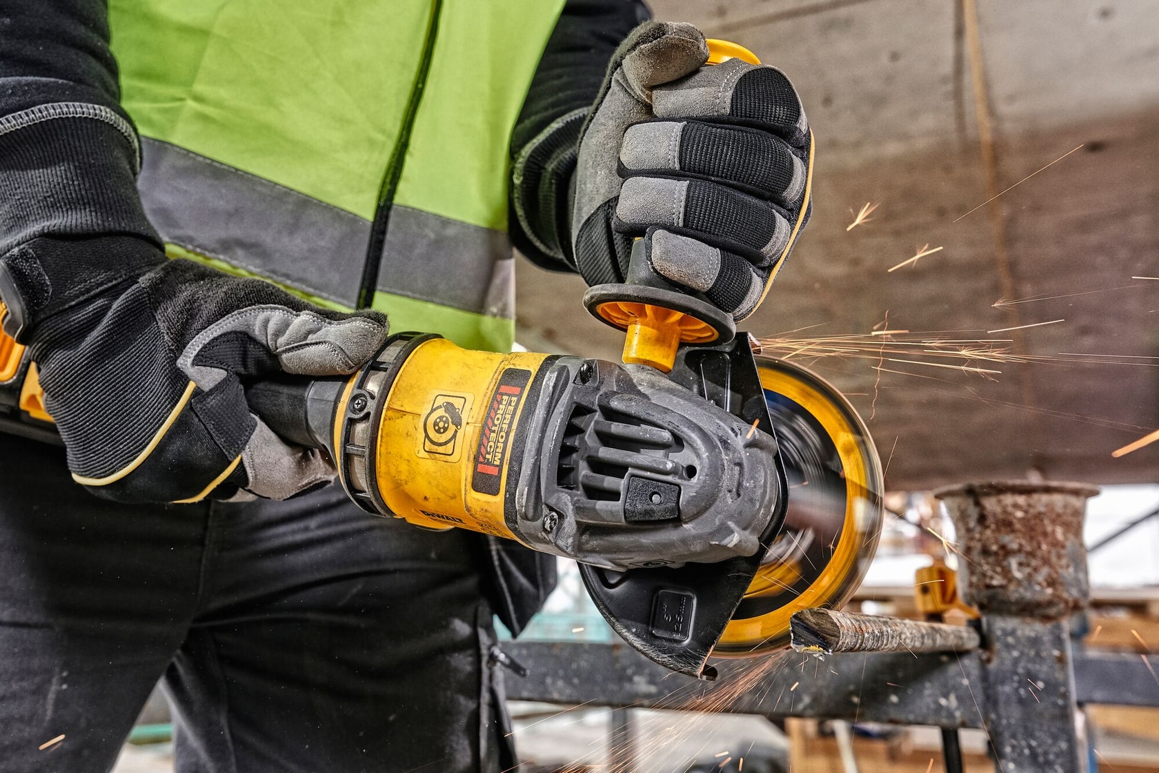 Una persona con guantes de seguridad y chaleco de alta visibilidad está usando una amoladora angular amarilla para cortar una barra de metal, generando chispas en un entorno de construcción.