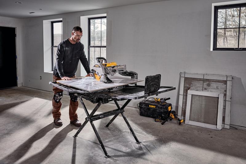 Una persona está al lado de una sierra para azulejos DEWALT D36000S montada en su soporte en una habitación en proceso de construcción. La sierra está sobre un soporte plegable negro y hay herramientas y una bolsa de herramientas DEWALT en el suelo cerca. La habitación tiene paredes sin terminar, ventanas y materiales de construcción apoyados contra la pared.