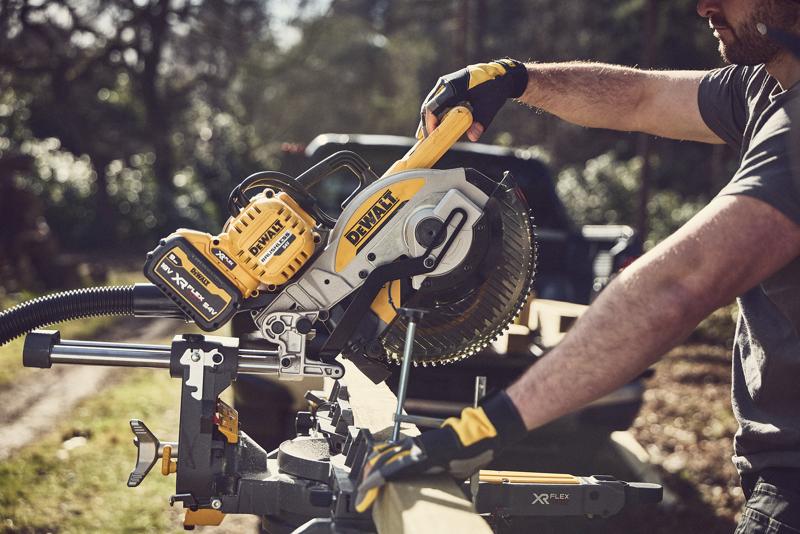 Una persona con guantes utiliza una sierra ingletadora inalámbrica DEWALT al aire libre para cortar un trozo de madera. La sierra tiene un diseño amarillo y negro con la marca DEWALT XR FLEXVOLT.
