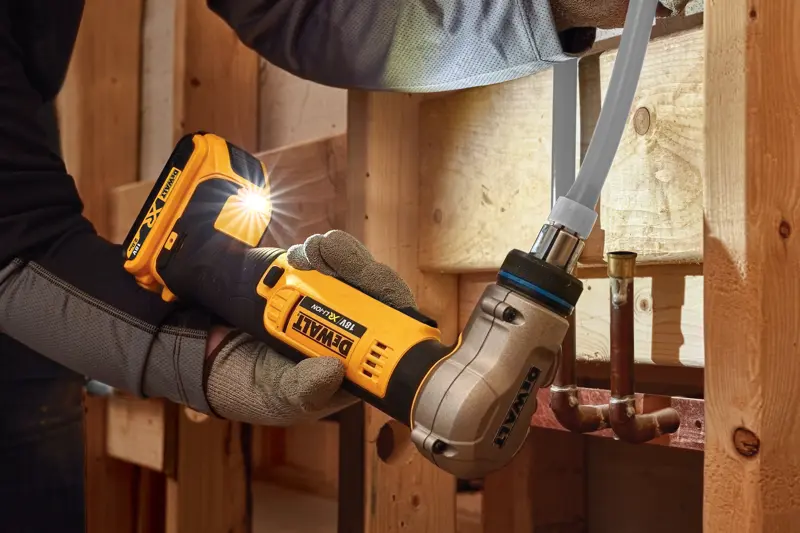 Una persona con guantes de trabajo utiliza una herramienta de engaste de tubos inalámbrica DEWALT DCE400D2 18V para conectar una tubería dentro de una estructura de madera.