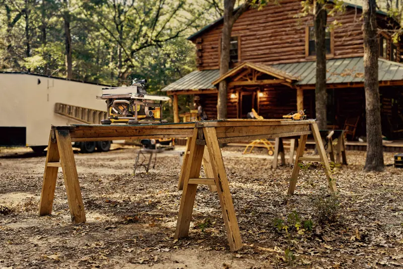 Una sierra de mesa DEWALT colocada en un banco de trabajo de madera formado por dos caballetes al aire libre, con una cabaña de troncos y árboles al fondo.