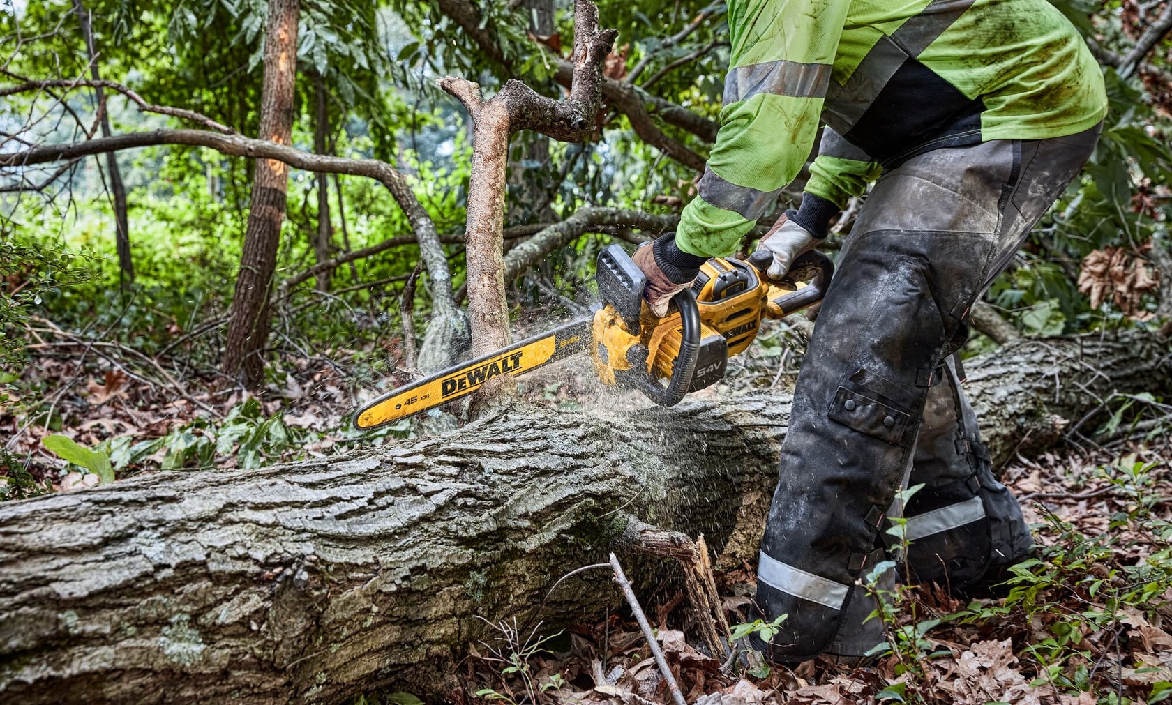 Una persona con equipo de protección utiliza una motosierra inalámbrica DEWALT para cortar un gran tronco caído en una zona boscosa.
