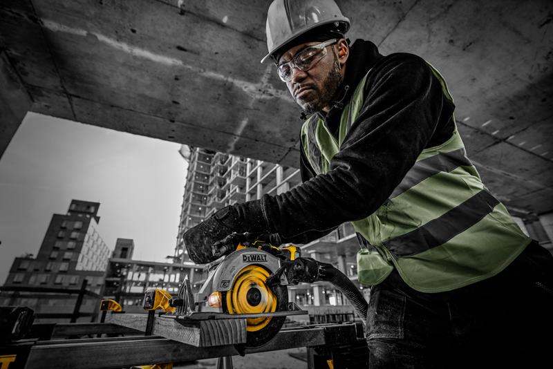 Un trabajador de la construcción con chaleco de seguridad y casco opera una sierra circular DEWALT (modelo DCS565NT) para cortar una tabla de madera en un sitio de construcción. Al fondo se observan estructuras de concreto y edificios en construcción.