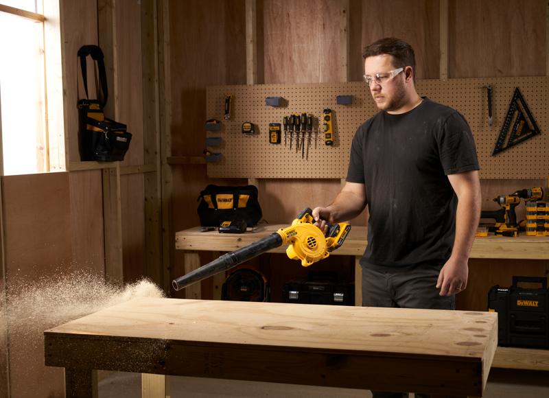 Una persona usa un soplador inalámbrico amarillo DEWALT para limpiar el aserrín de un banco de trabajo de madera en un taller. Varias herramientas y bolsas de herramientas están organizadas en la pared del fondo.