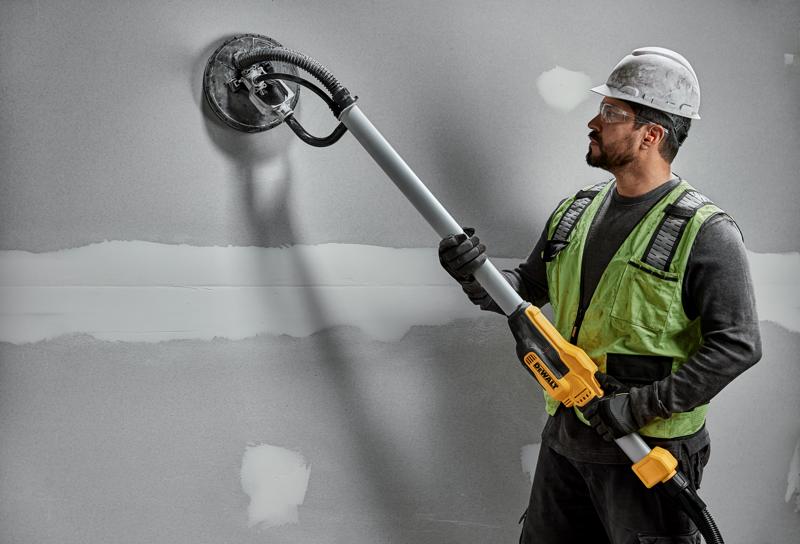 Un trabajador de la construcción con casco y chaleco de seguridad utiliza una lijadora de paneles de yeso DEWALT DWE7800 con mango telescópico para lijar una pared gris.