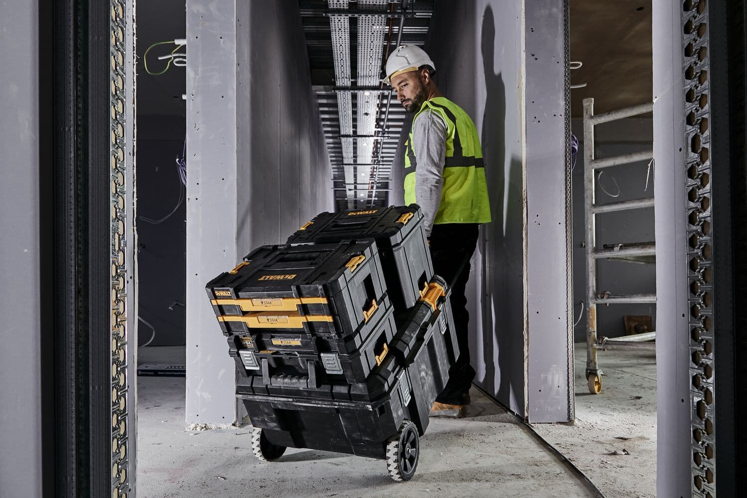 Una persona con chaleco de seguridad y casco tira de una pila de cajas de herramientas DEWALT con ruedas por un pasillo de obra.