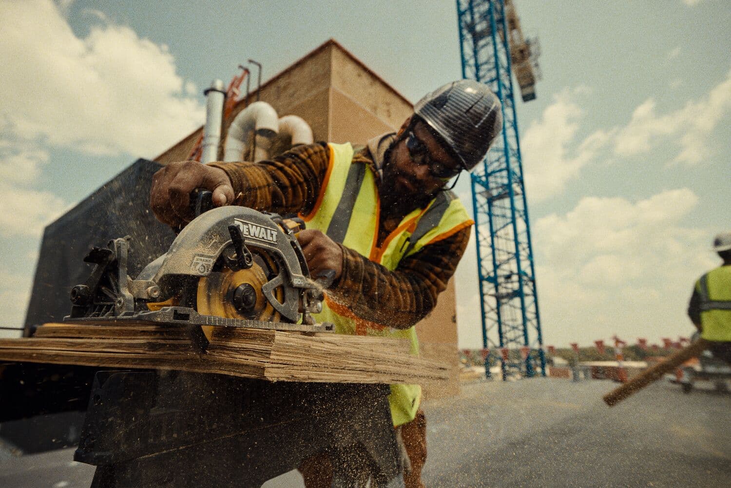 A construction worker wearing a safety vest and helmet is using a DEWALT circular saw to cut wooden boards at a construction site. Sawdust is visible as the tool operates, with another worker and a crane in the background.