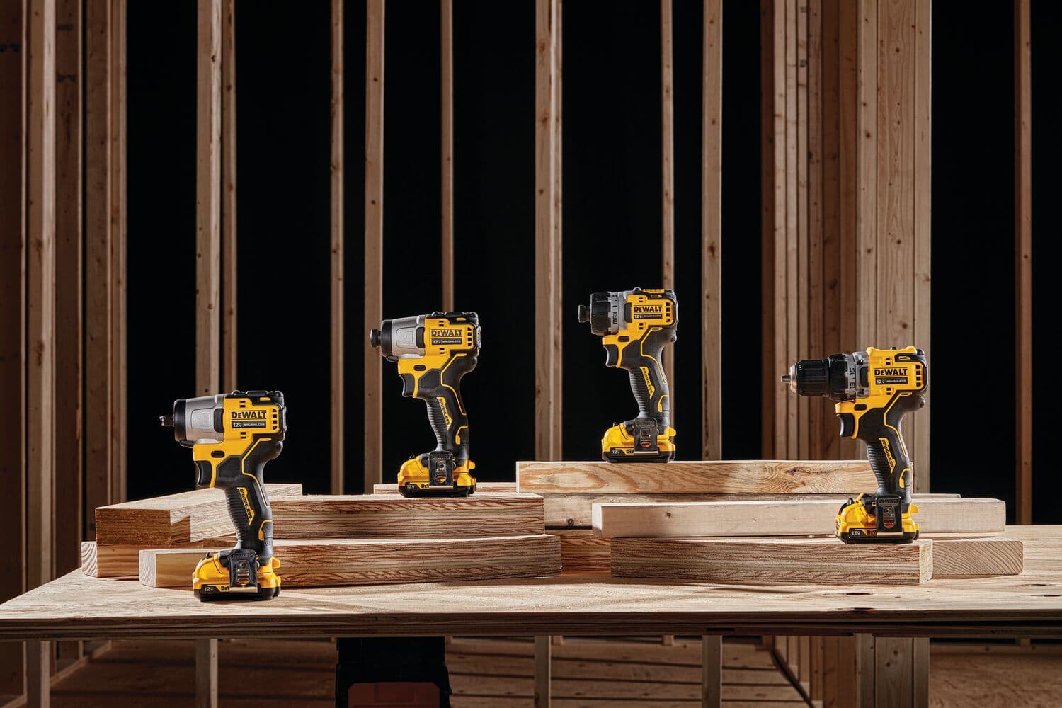 Cuatro atornilladores de impacto inalámbricos DEWALT están colocados sobre bloques de madera en una mesa de trabajo, con una estructura de marco de madera en el fondo. Las herramientas son principalmente de color amarillo y negro y están exhibidas en un entorno de construcción.
