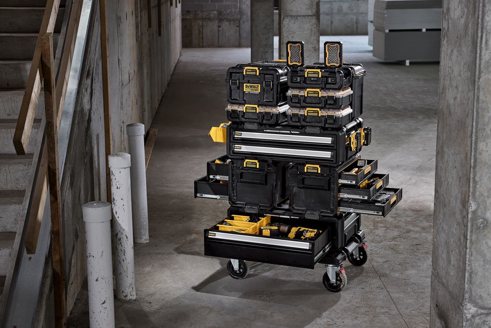 A DEWALT mobile tool storage cart with multiple stacked black toolboxes and drawers, shown open to display organized tools and accessories. The cart is on wheels and situated in an unfinished industrial or construction site.