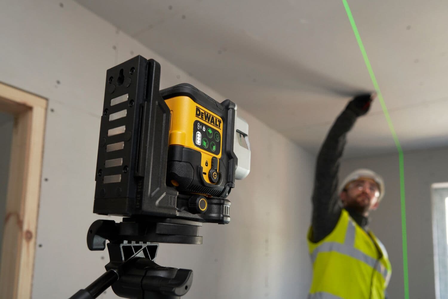 Un nivel láser DEWALT montado en un trípode proyecta una línea verde brillante en el techo de una zona de construcción. Una persona con un chaleco reflectante marca o mide a lo largo de la línea.