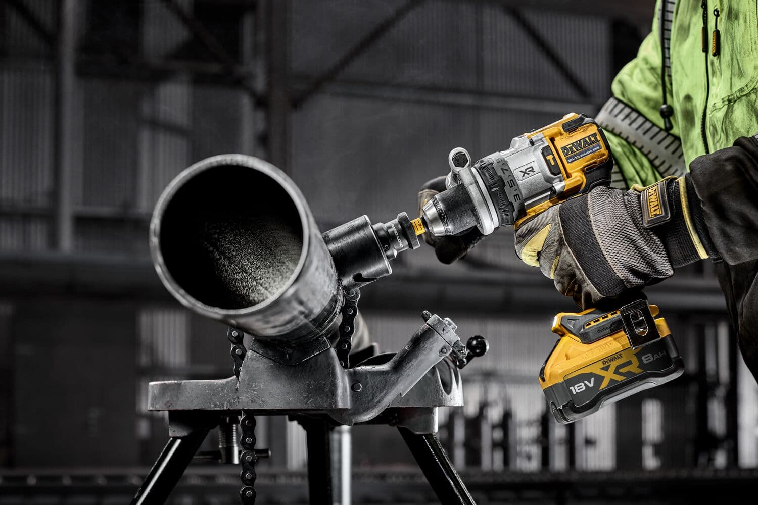A person wearing safety gloves and a green jacket is using a DEWALT cordless drill to cut or bore into a large metal pipe fixed on a stand in an industrial setting.
