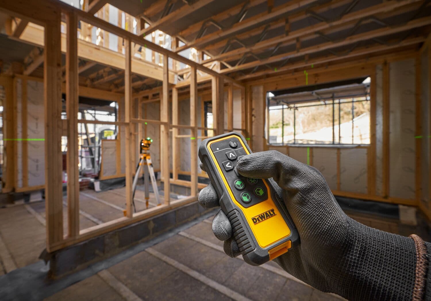 Una mano enguantada sostiene un dispositivo de control remoto DEWALT en primer plano, con un nivel láser DEWALT colocado sobre un trípode en un sitio de construcción de estructura de madera en el fondo. Líneas láser verdes son visibles en el lugar para ayudar con la alineación y las mediciones.