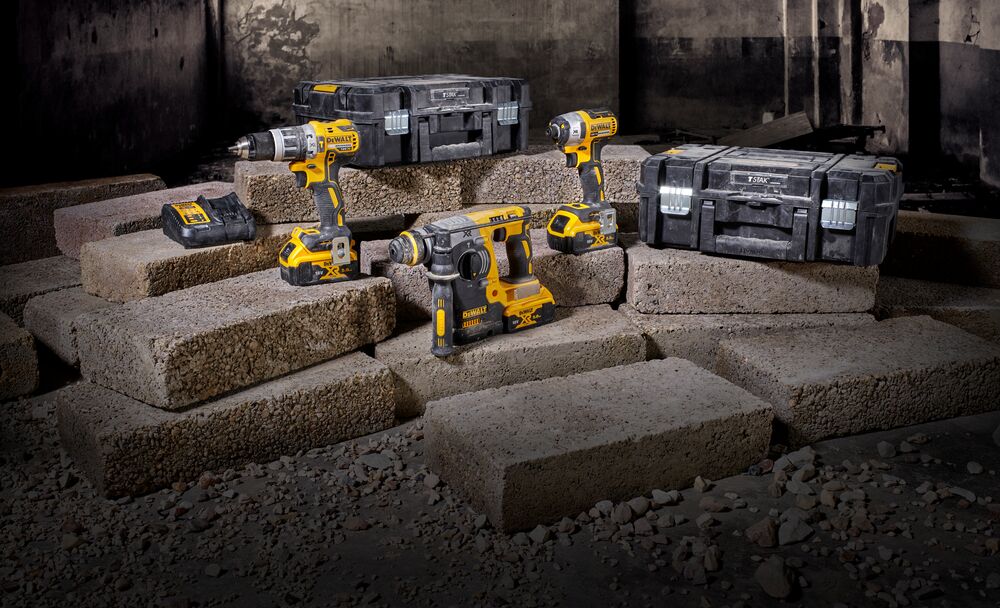 A set of DEWALT cordless power tools is displayed on concrete blocks. The set includes a cordless drill, an impact driver, a rotary hammer, two sturdy black toolboxes, and a battery charger. The tools feature a yellow and black color scheme.
