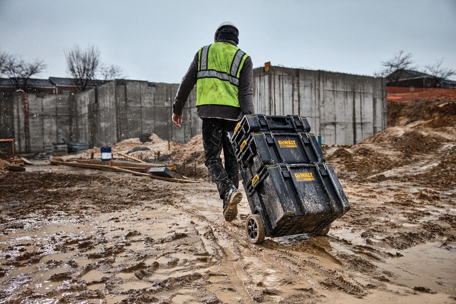 Una persona con chaleco de seguridad y casco está arrastrando una pila de cajas de herramientas DEWALT con ruedas a través de un sitio de construcción embarrado.