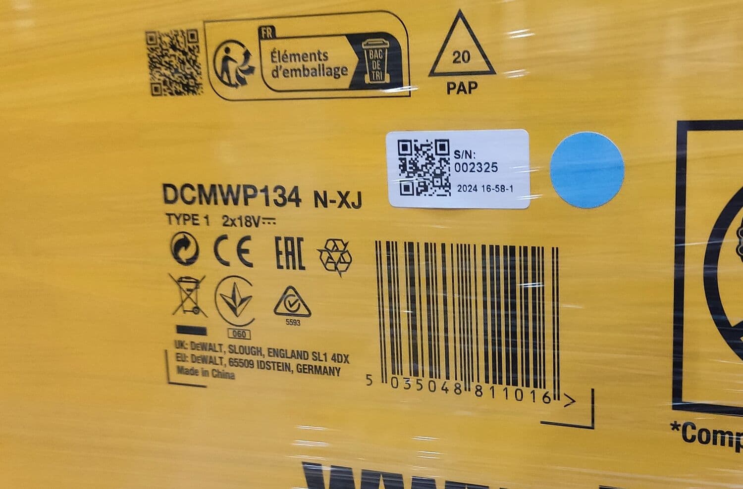 Close-up of a DEWALT product packaging showing various markings and labels. The packaging includes QR codes, recycling symbols, regulatory marks (CE, EAC), a warning symbol, and a barcode (5035048810116). The model number DCMWP134 N-XJ, type 1 2x18V, and serial number 002325 are visible. Details of the UK and EU addresses and 'Made in China' are printed. The packaging is partially covered with transparent plastic wrap.
