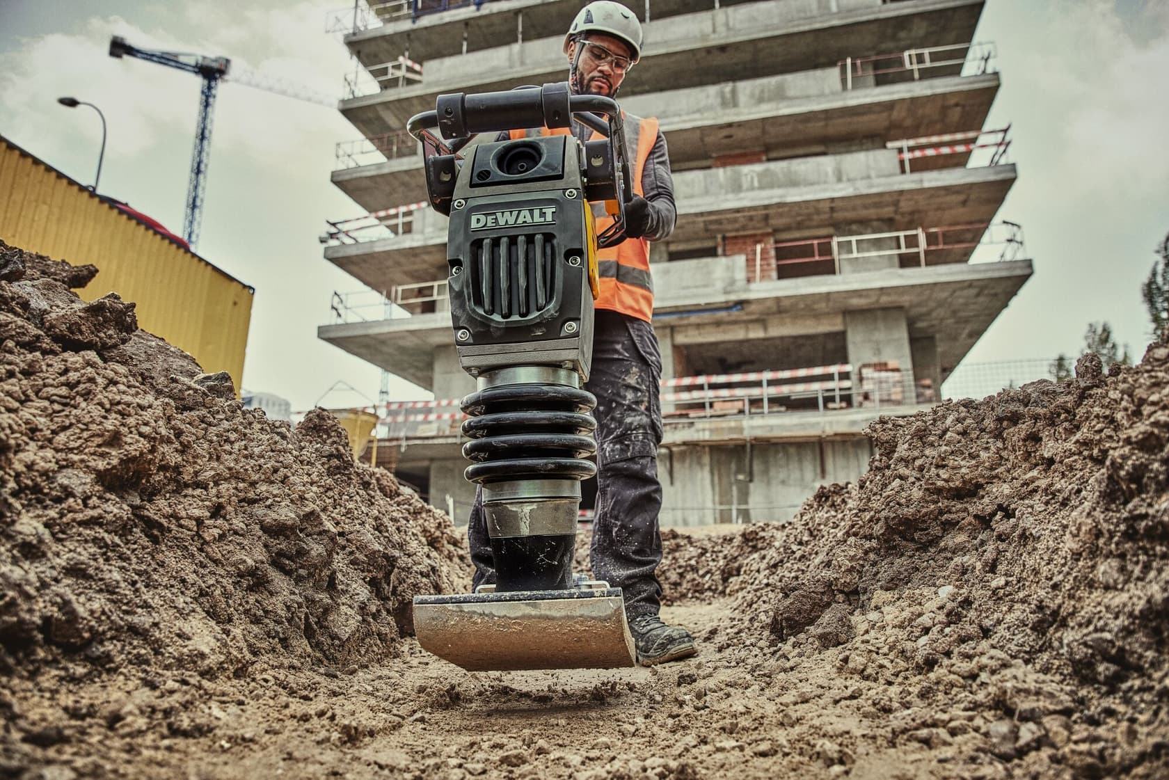 Un trabajador de construcción con casco y chaleco de seguridad opera una compactadora DEWALT sobre un camino de tierra en un sitio de construcción, con un edificio y una grúa visibles al fondo.