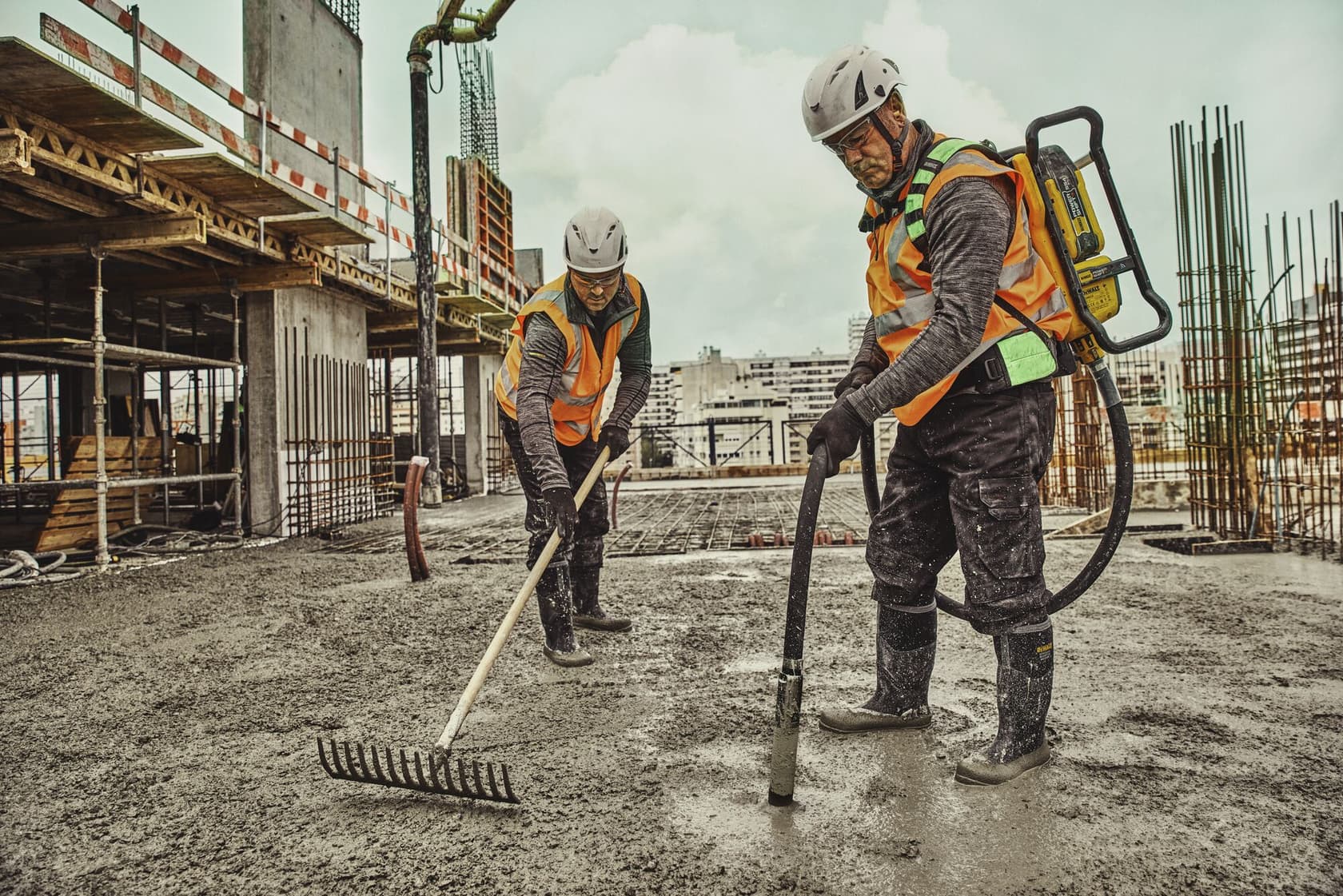 Dos trabajadores de la construcción con cascos de seguridad, guantes y chalecos naranjas están trabajando en una obra. Uno sostiene un rastrillo para nivelar el concreto, mientras que el otro utiliza un vibrador de concreto DEWALT tipo mochila. En el fondo se observa una estructura parcialmente construida con refuerzos de acero.
