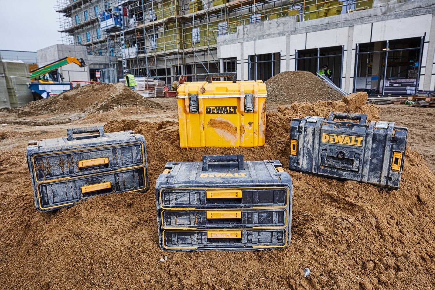 Four DEWALT branded toolboxes and organizers, in yellow and black, shown on a construction site with dirt and a building under construction in the background.