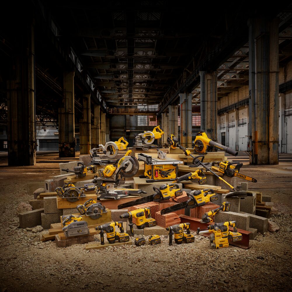 Una variedad de herramientas eléctricas DEWALT en amarillo y negro, incluyendo taladros, sierras, sopladores y una sierra de mesa, están exhibidas sobre tablas de madera, ladrillos y bloques de concreto apilados dentro de un gran almacén industrial.