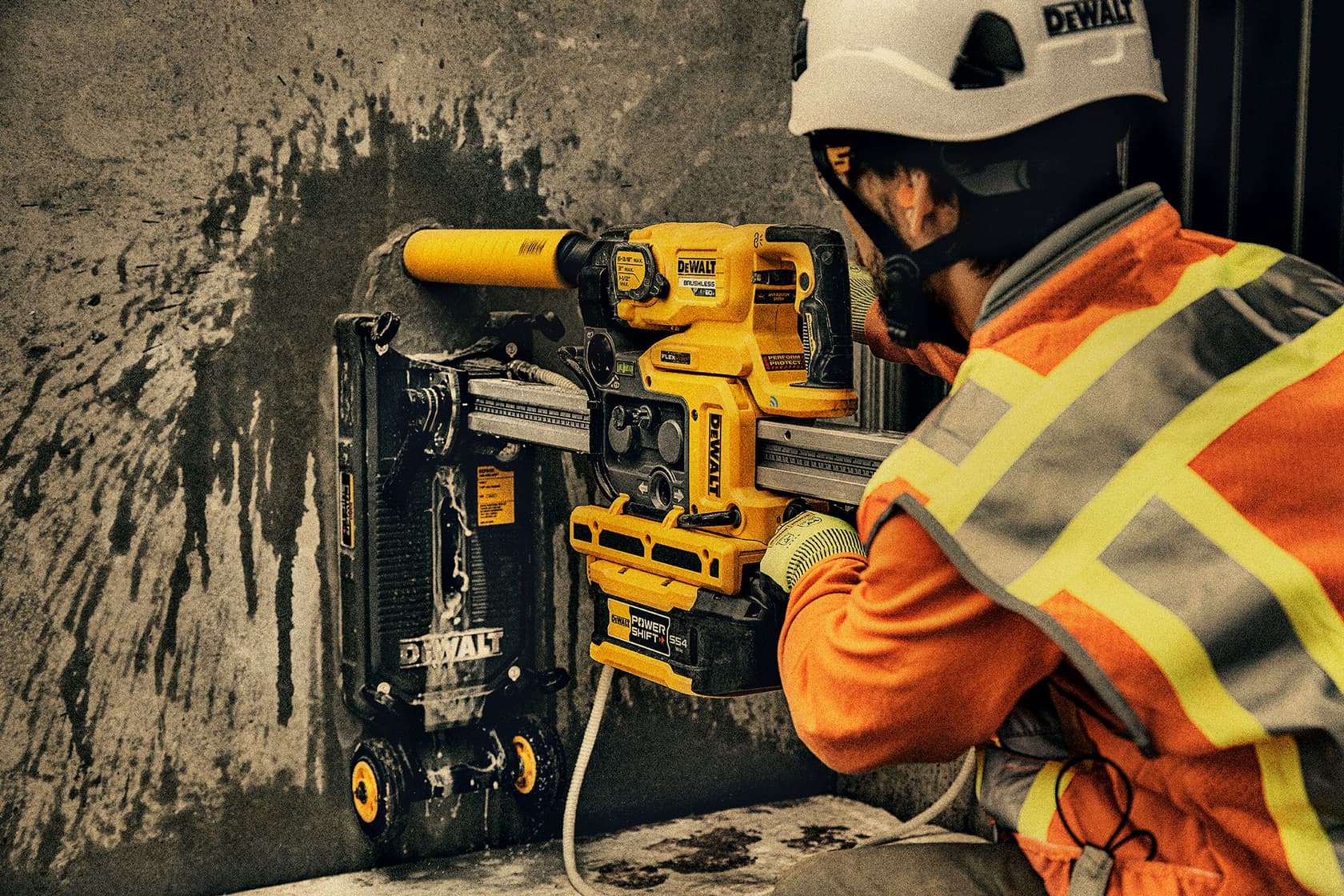 Un trabajador de la construcción con un chaleco de seguridad naranja y casco utiliza una herramienta eléctrica DEWALT montada en una pared de concreto.