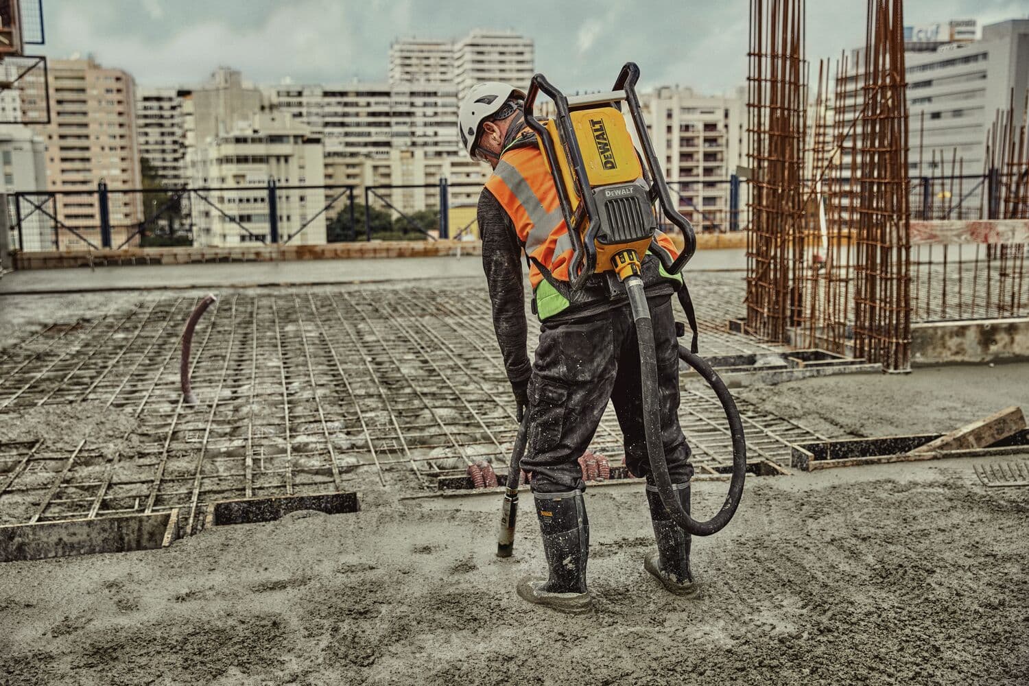Un trabajador de la construcción con equipo de seguridad y un vibrador de concreto DEWALT tipo mochila está trabajando en una losa de concreto en un sitio de construcción con edificios altos al fondo.